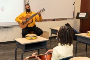 Indy STEAM Academy music teacher demonstrates how to play the guitar during music class. An Indy STEAM student watches and plays a guitar.