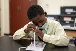 An Indy STEAM Academy student looks through a lens during science class. The student wears a yellow shirt and blue tie. 