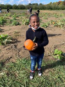 An Indy STEAM Academy student stands in a pumpkin patch and smiles while holding a pumpkin she picked.