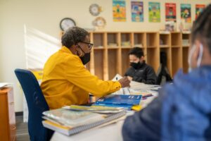 An Indy STEAM Academy teacher sits at a table with two students in small groups in a classroom. All are working quietly on a task.