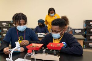 Two Indy STEAM Academy students work together in a science lab on measurement using weights and balances. Behind them, an Indy STEAM Academy teacher supports two students in their work. 