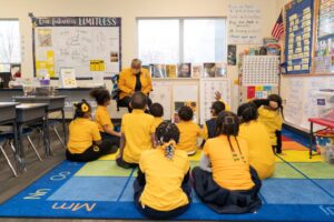 Indy STEAM Academy students in brightly colored yellow and blue uniforms sit together on a classroom carpet facing their teacher who is also wearing a yellow uniform shirt. 