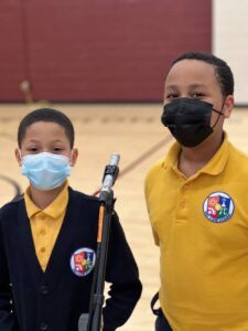 Two Indy STEAM Academy students in blue and yellow uniforms smile while speaking together at a microphone in the school gymnasium. Both students are wearing face masks.