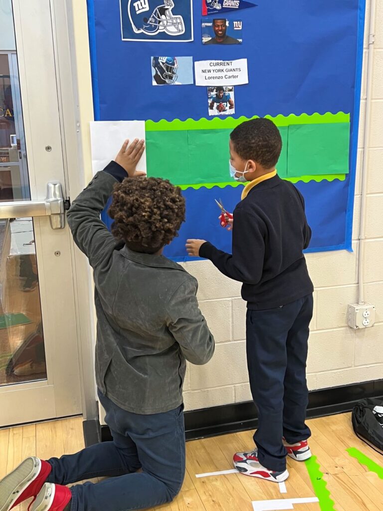 Two Indy STEAM Academy students wearing blue uniforms work together to create a bulletin board project.