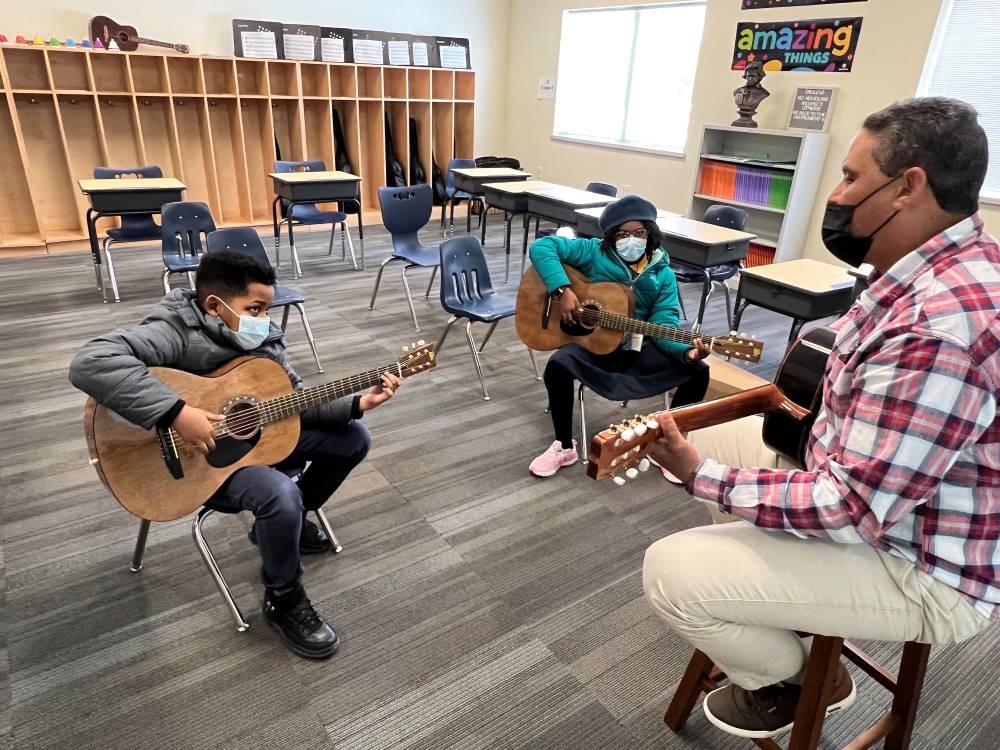 An Indy STEAM Academy instructor guides two students through a guitar lesson. They are seated in a music room at the school. Each person is holding a guitar.