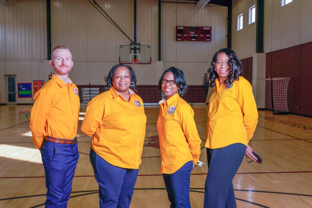 the 4 members of the Indy STEAM Academy leadership team stand together for a photograph in the school gymnasium. They are wearing yellow button down shirts and navy blue pants, the school uniform.
